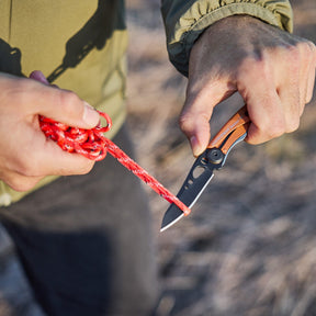 Hand using an Aurora Leatherman Skeletool KB to cut red rope outdoors